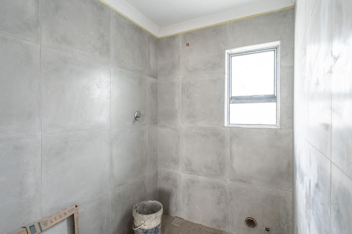 An unfinished bathroom space featuring large format grey wall tiles, a window, and exposed plumbing connections, currently in a state of renovation.
