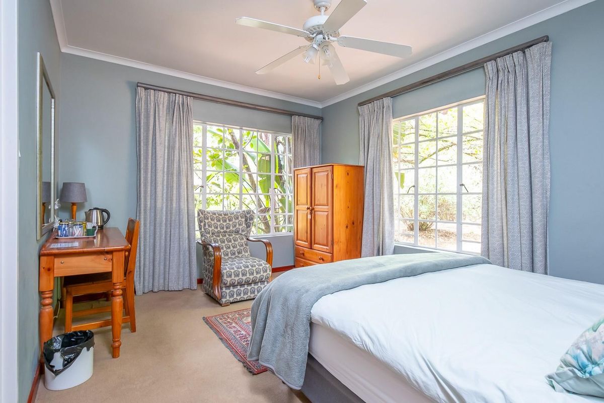 A well-lit bedroom featuring large windows with garden views, neutral carpeting, and a ceiling fan.