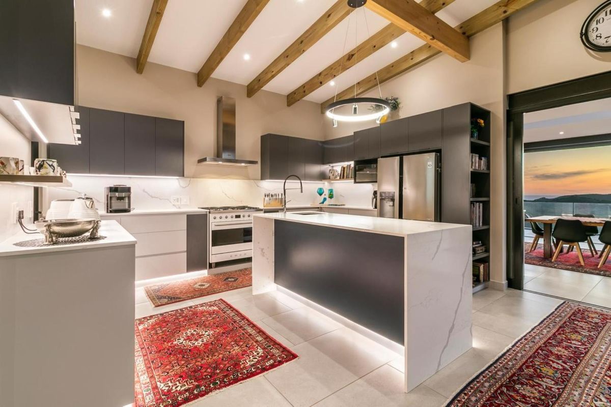 Modern kitchen featuring a central island with stone countertops, dark cabinetry, exposed timber ceiling beams, and integrated lighting, flowing into an outdoor patio area.