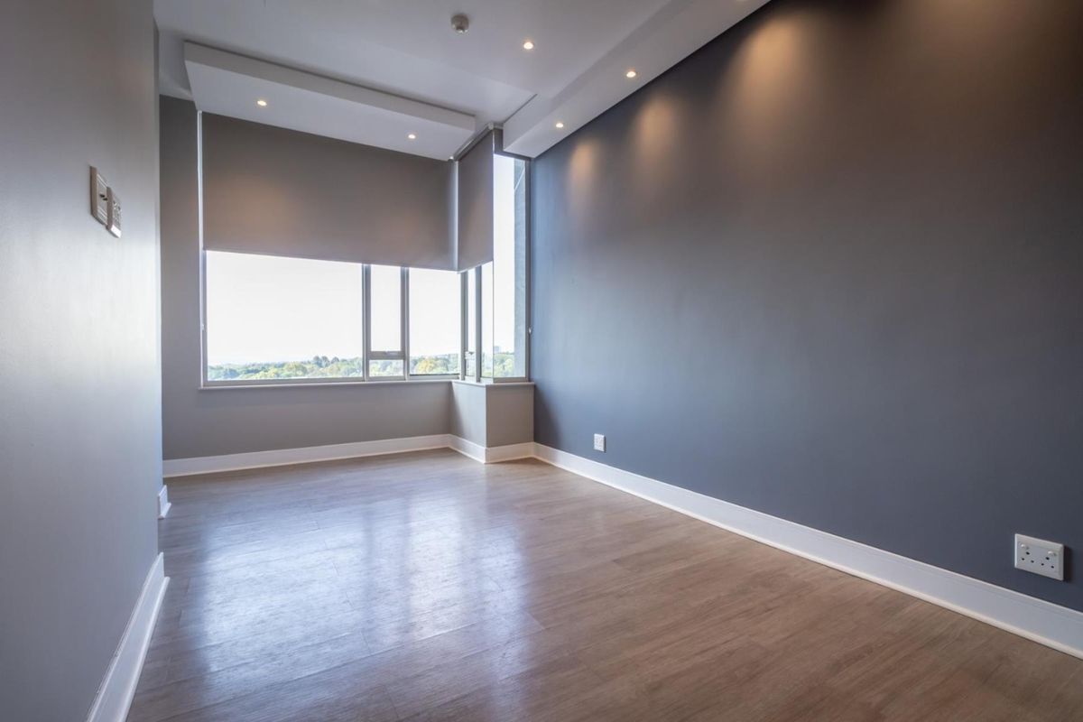A bright, empty bedroom featuring modern grey accent walls, recessed ceiling lighting, and large windows with roller blinds, finished with wood-look flooring.