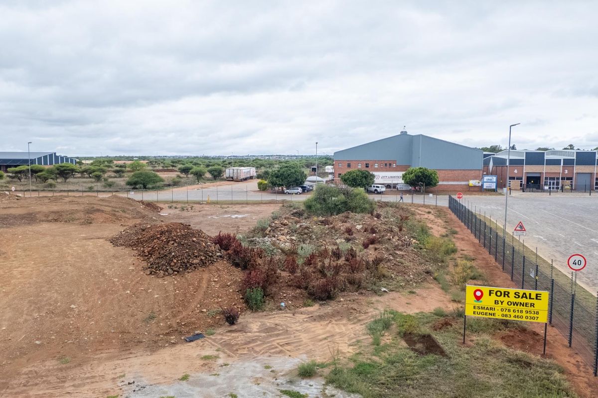 An elevated view of a vacant residential plot featuring natural terrain, perimeter fencing, and adjacent commercial infrastructure.