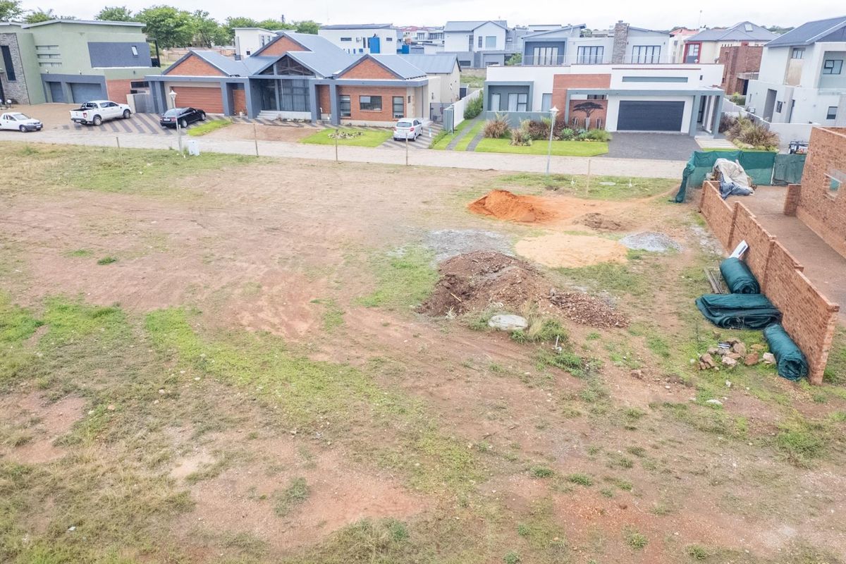 An elevated view of an undeveloped residential plot featuring raw earth, sparse vegetation, and surrounding modern housing developments.