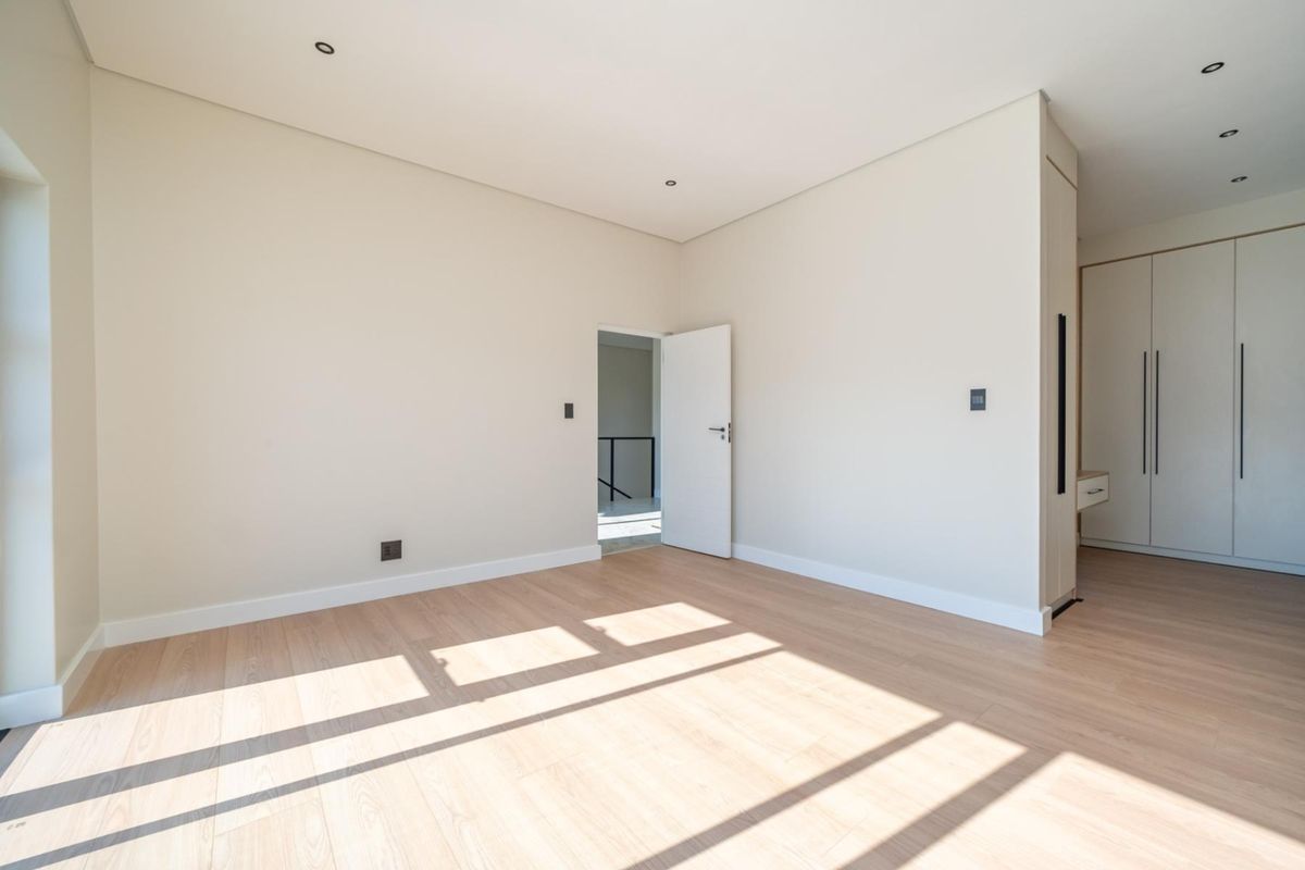A bright, modern bedroom featuring light wood-look flooring, neutral walls, recessed ceiling lighting, and built-in floor-to-ceiling cabinetry.