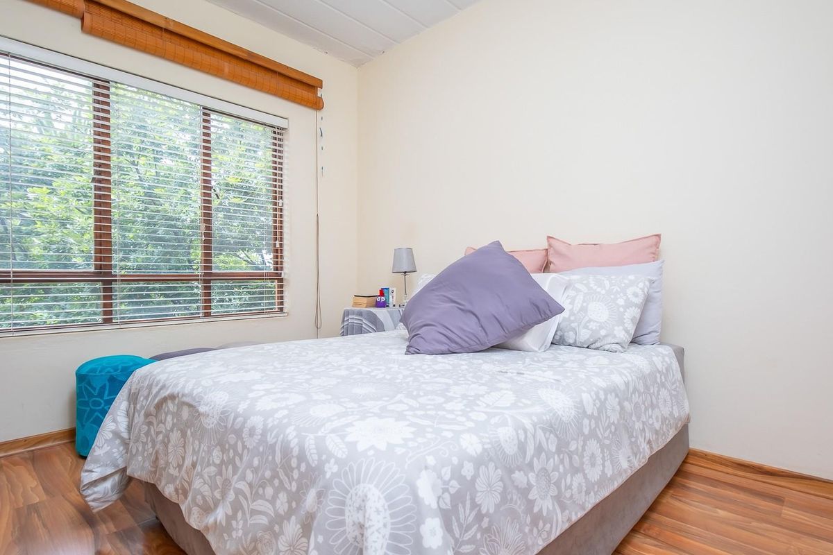 A bedroom featuring wood-look laminate flooring and a large window with fitted blinds, providing natural light and a view of the surrounding greenery.