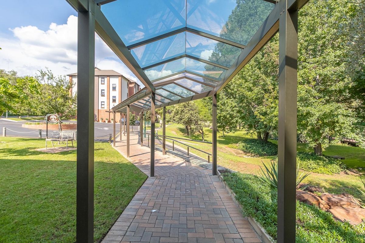 A well-maintained paved walkway featuring a modern glass-roofed structure, set within a lush landscaped garden area of the apartment complex.