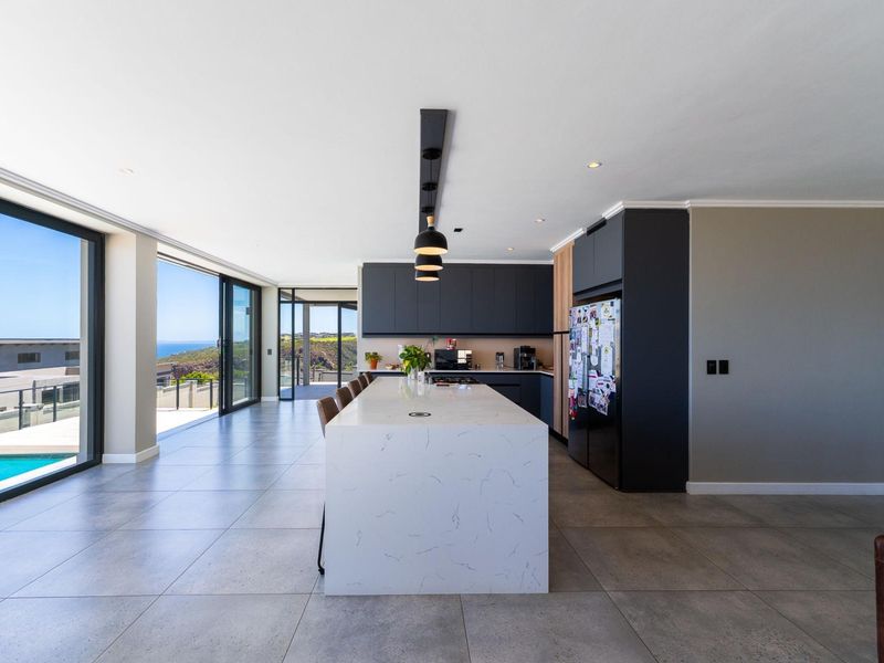 Modern open-plan kitchen featuring a large stone-topped island, sleek dark cabinetry, and expansive sliding glass doors opening to an outdoor area with coastal views.
