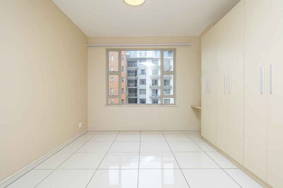 A bright, empty bedroom featuring large white floor tiles, neutral walls, and a wall-to-wall built-in wardrobe with integrated shelving.