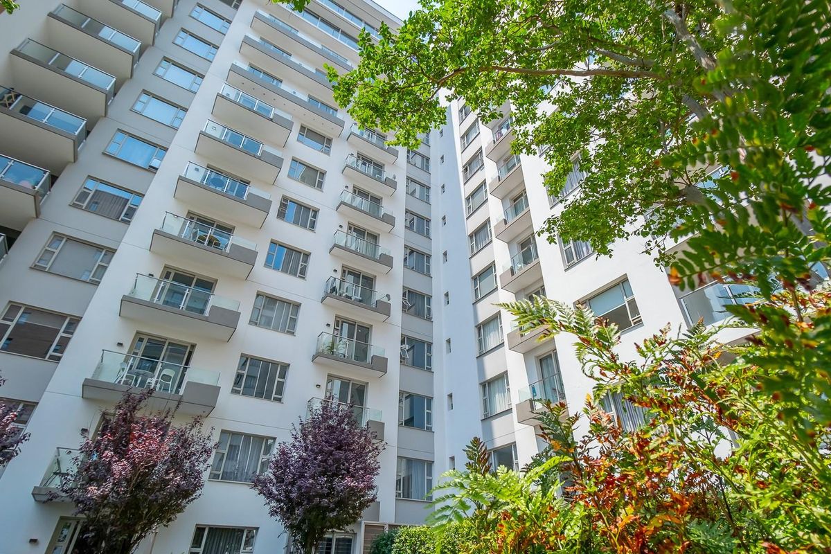 Low-angle view of a modern multi-story apartment building featuring numerous private balconies and large windows, surrounded by landscaped greenery.