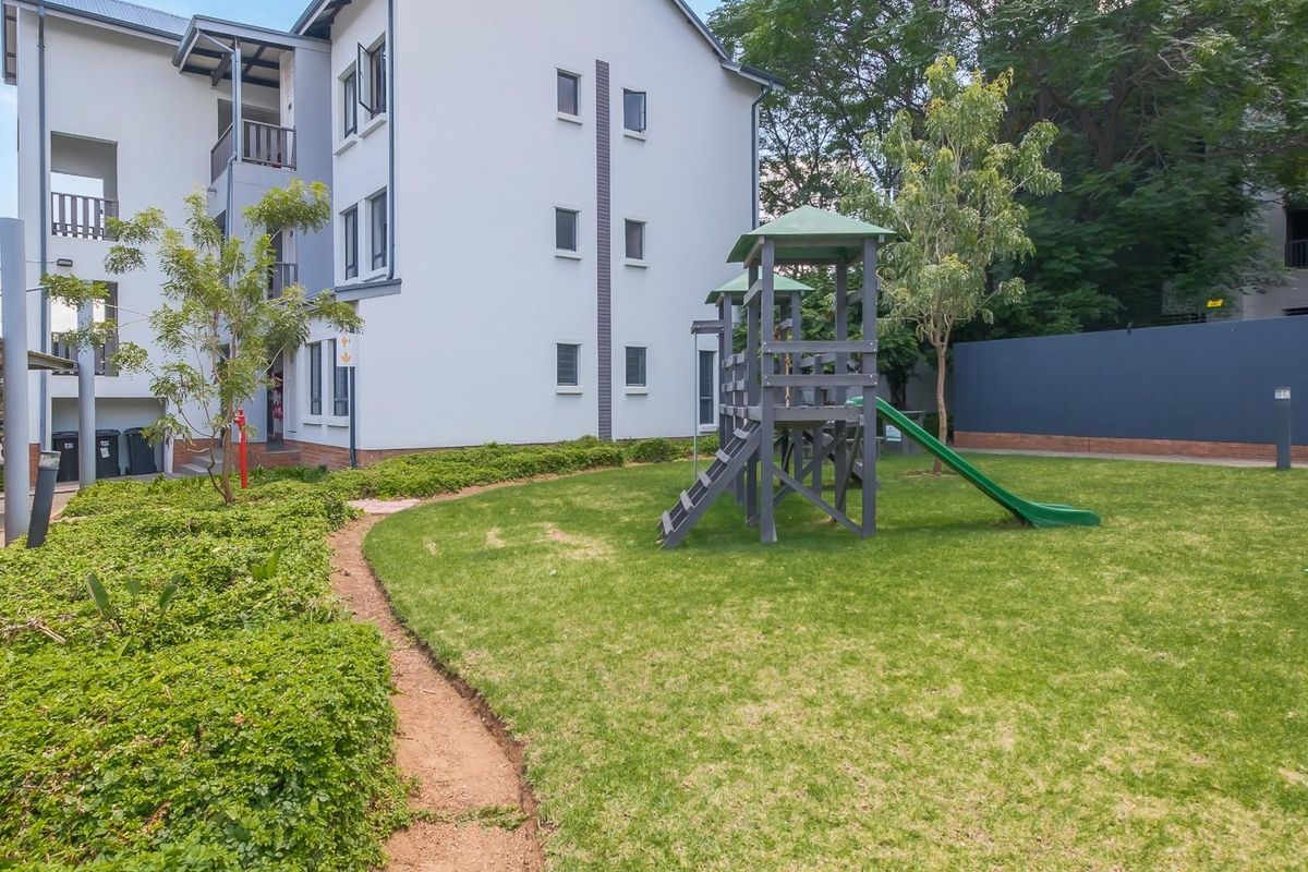 A well-maintained communal garden area featuring a children's playground set, manicured lawn, and paved pathways adjacent to the apartment building.