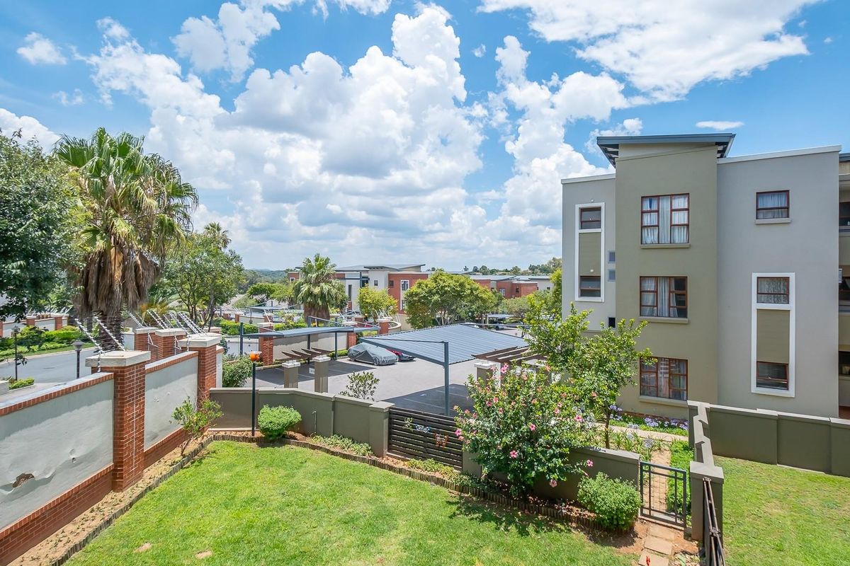 Exterior view of the apartment complex featuring a landscaped garden, perimeter brick wall, and covered parking area under a clear blue sky.
