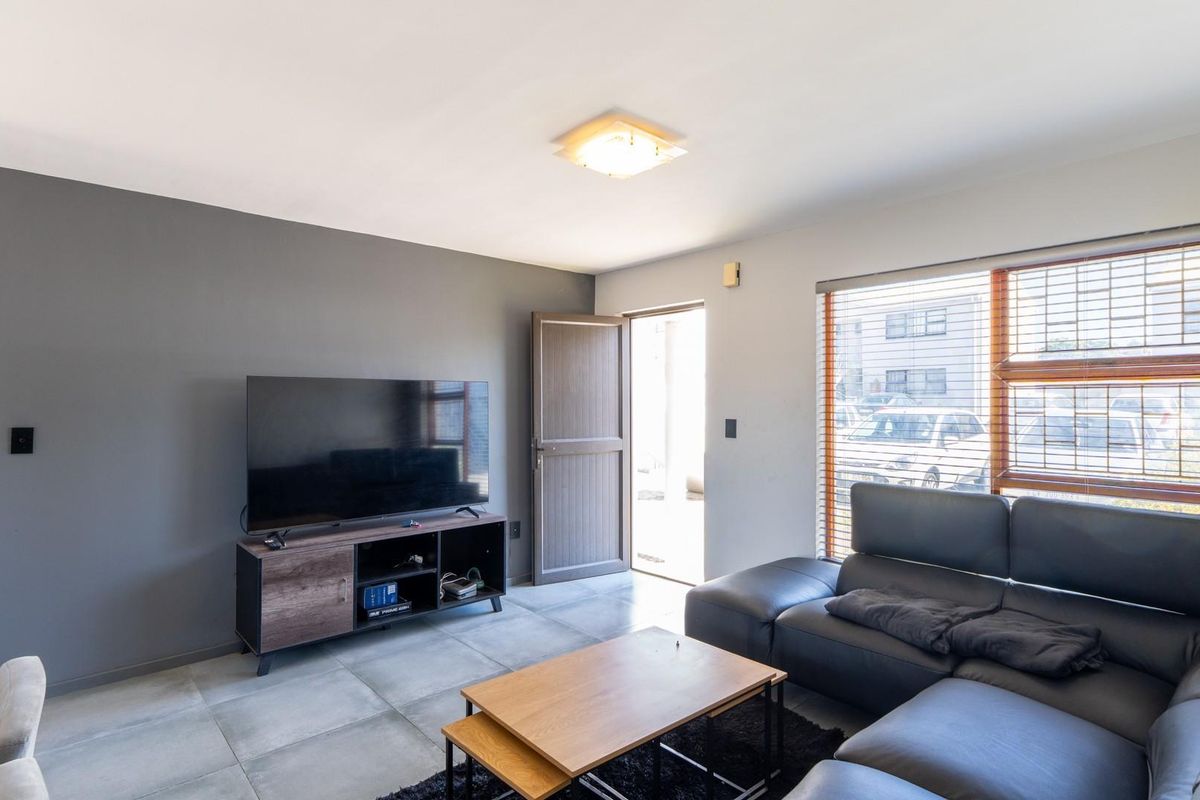 A well-lit living room featuring large floor tiles, neutral grey accent wall, and natural light through venetian blinds.