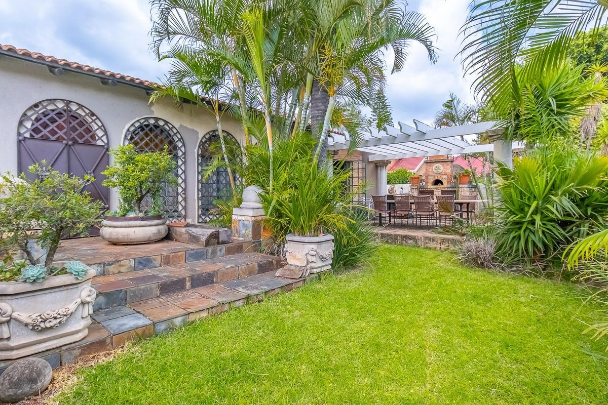 A landscaped rear garden featuring a stone-paved patio, a pergola-covered outdoor entertainment area with a built-in brick fireplace, and mature tropical plantings.