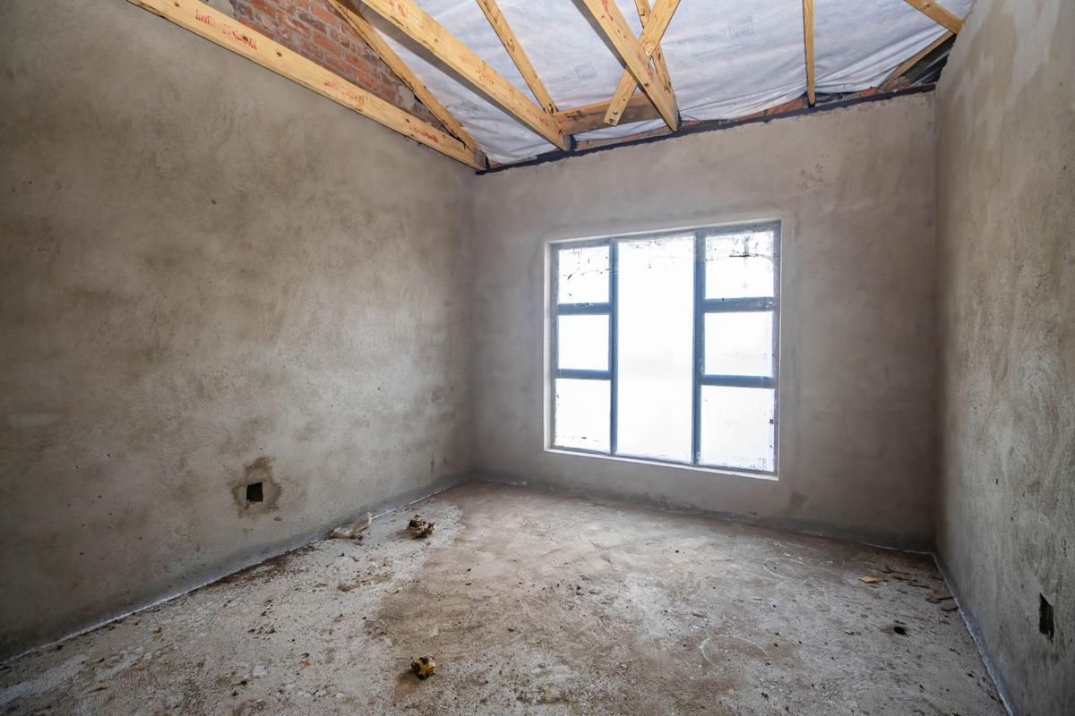 An unfinished bedroom space featuring exposed roof trusses, raw plaster walls, and a large window, currently in a shell condition awaiting final finishes.