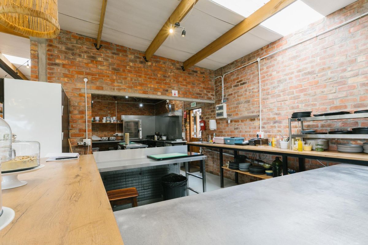A rustic industrial-style kitchen featuring exposed brick walls, timber ceiling beams, stainless steel prep counters, and a functional commercial-grade layout.