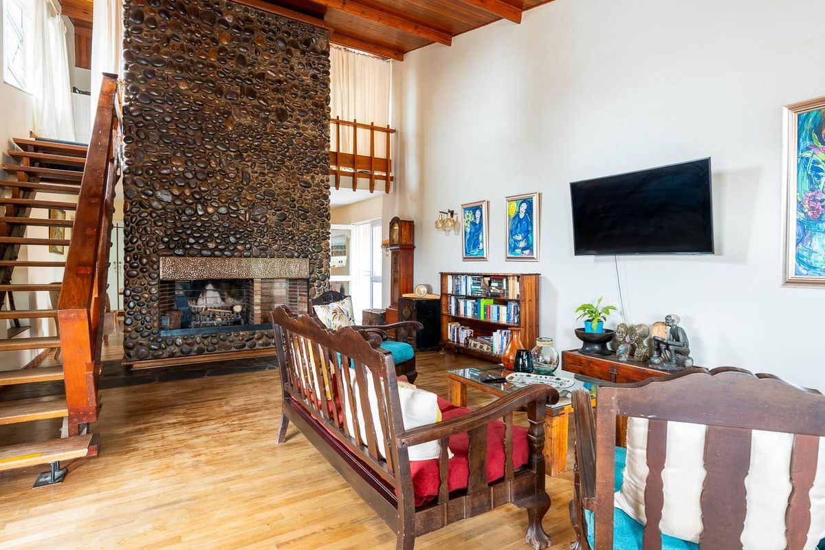 Spacious living room featuring a prominent floor-to-ceiling river stone fireplace, exposed timber ceiling beams, and a wooden staircase leading to an upper mezzanine level.