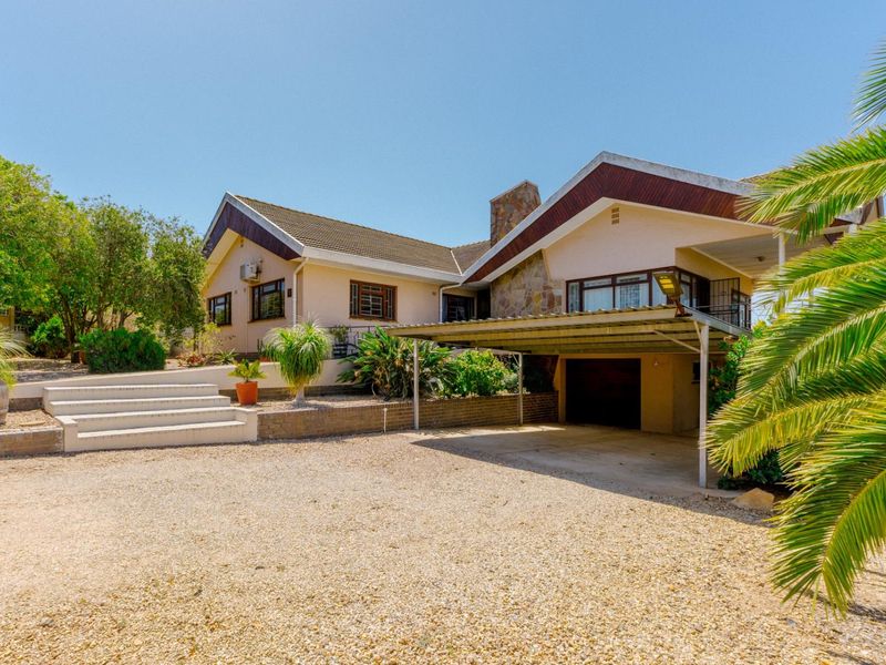 Front exterior view of a residential house featuring a gravel driveway, covered carport, stone-clad chimney, and landscaped garden with concrete steps.