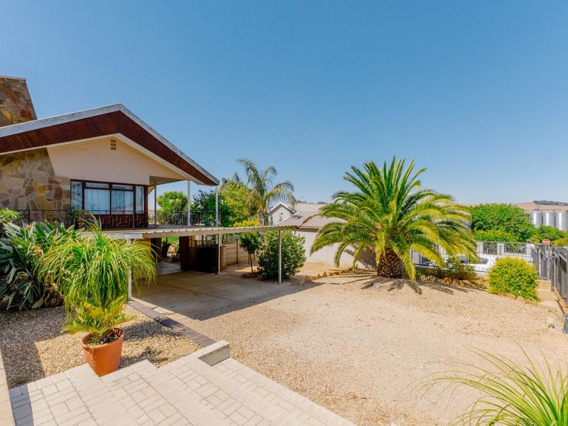 Front exterior view of the house featuring a covered carport, stone-clad facade, and a landscaped garden area with mature palm trees.