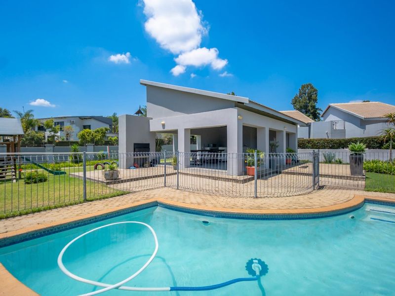 A well-maintained outdoor pool area featuring a paved deck, metal safety fencing, and a covered patio with a built-in braai.