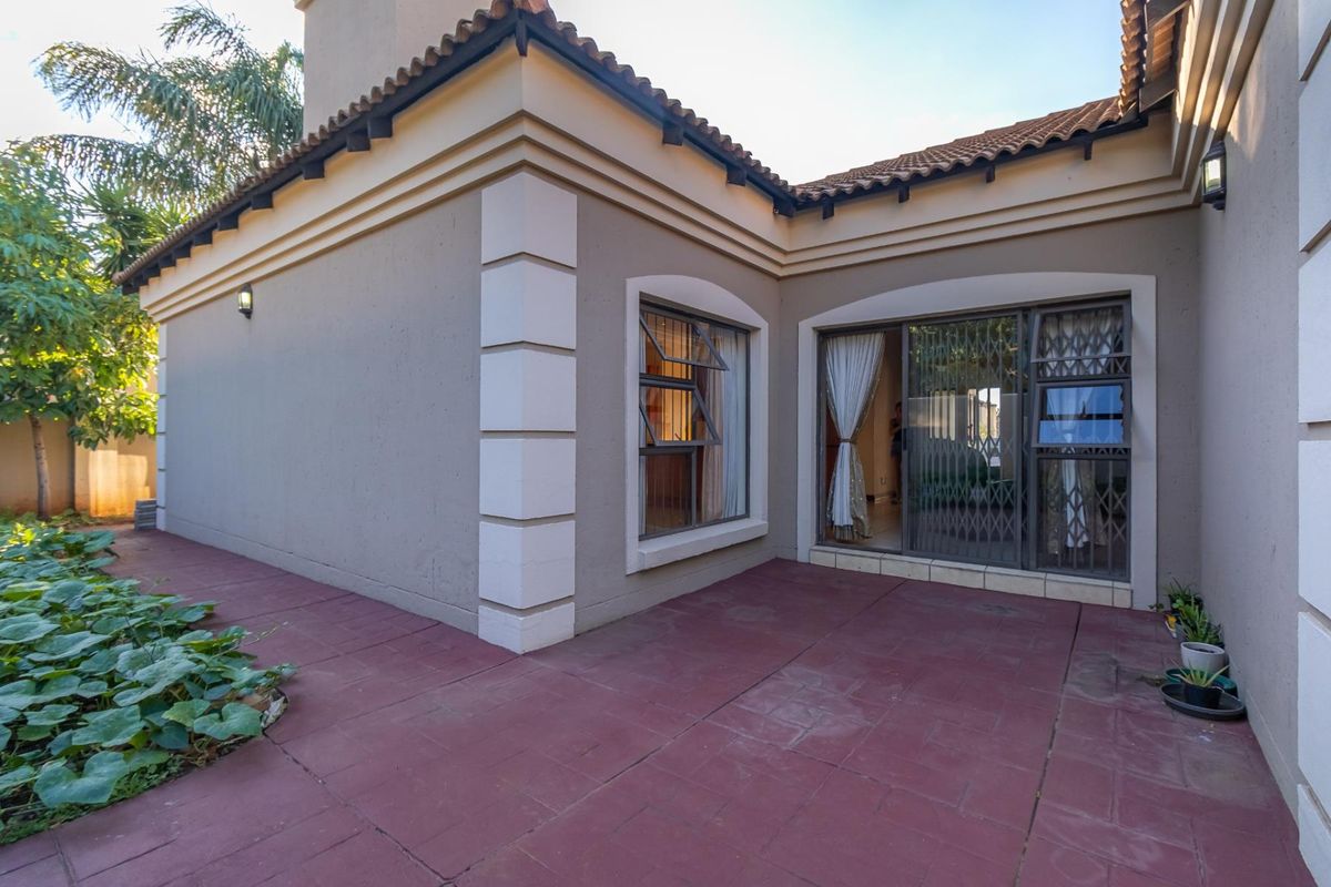 A rear exterior view featuring a paved patio area, sliding glass doors with security gates, and well-maintained exterior walls with terracotta roof detailing.