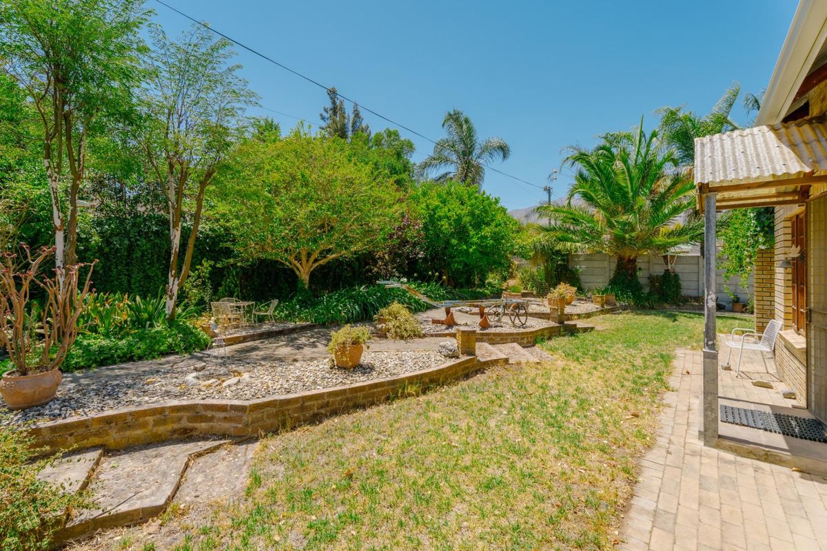 A spacious rear garden featuring tiered brick retaining walls, established trees, and a paved patio area adjacent to the house.