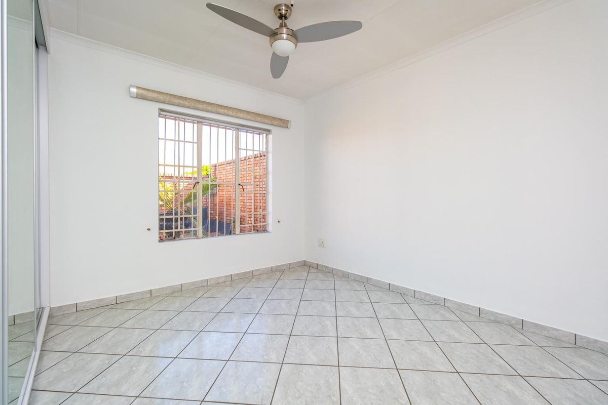 A bright, empty bedroom featuring tiled flooring, a ceiling fan, and a window fitted with security bars.
