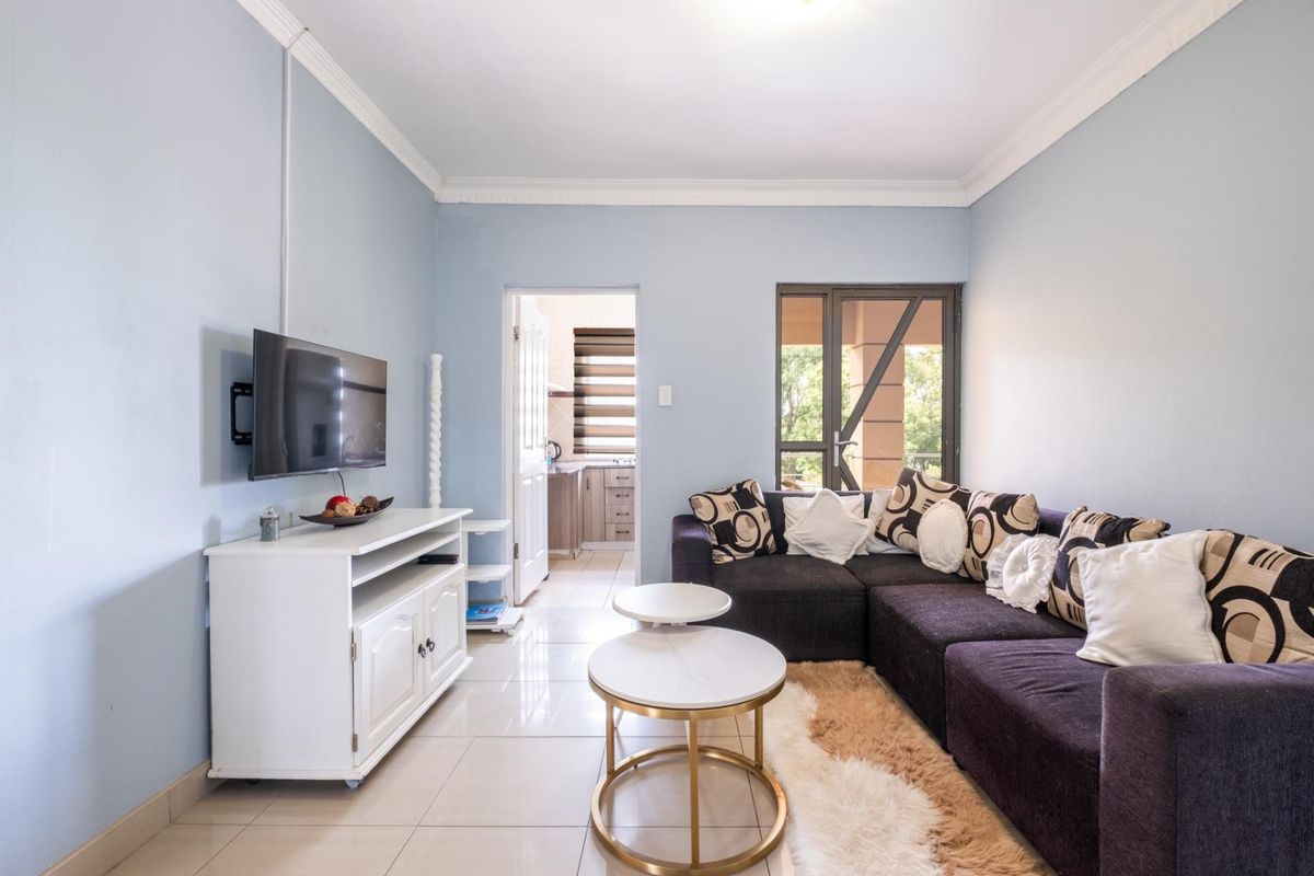 A bright living room featuring tiled flooring, crown molding, and direct access to an adjacent kitchen and outdoor balcony area.