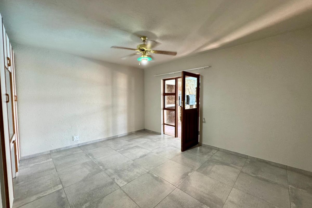 A bright, empty living room featuring large format grey floor tiles, a ceiling fan, and a wooden door leading to an outdoor area.