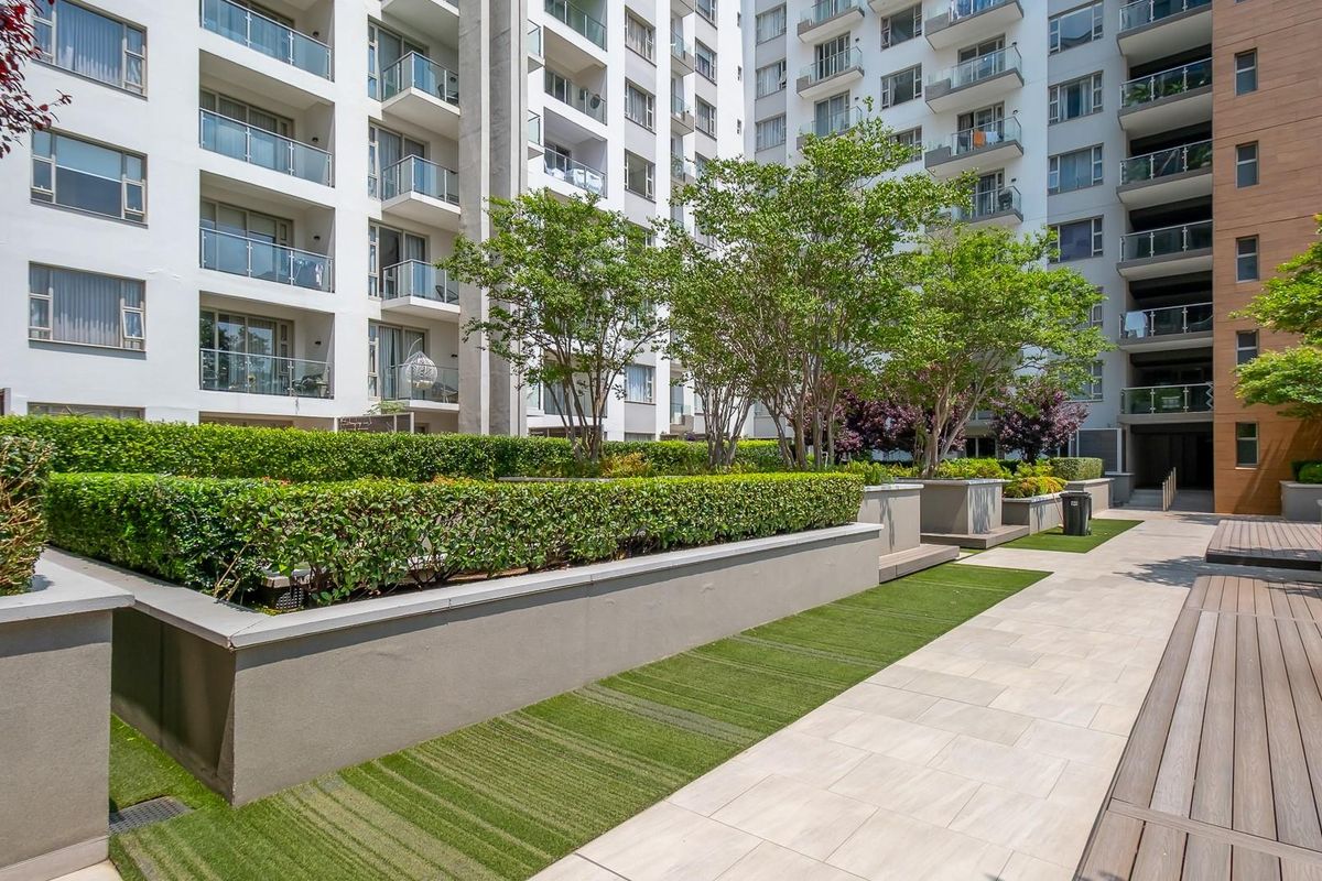A well-maintained communal garden area featuring raised planters, manicured hedges, paved walkways, and timber decking, situated between apartment building blocks.