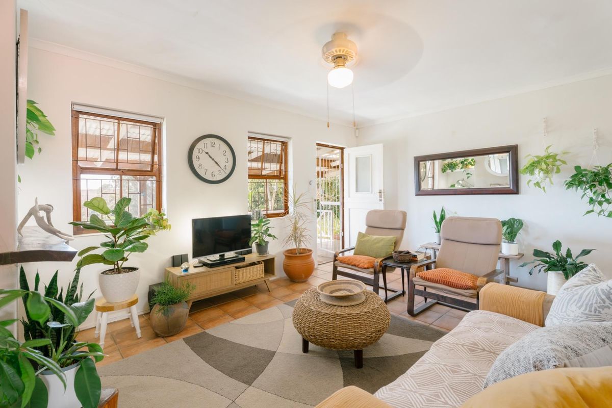 A bright living room featuring terracotta floor tiles, a ceiling fan, and wooden window frames with security bars, providing access to an outdoor area.