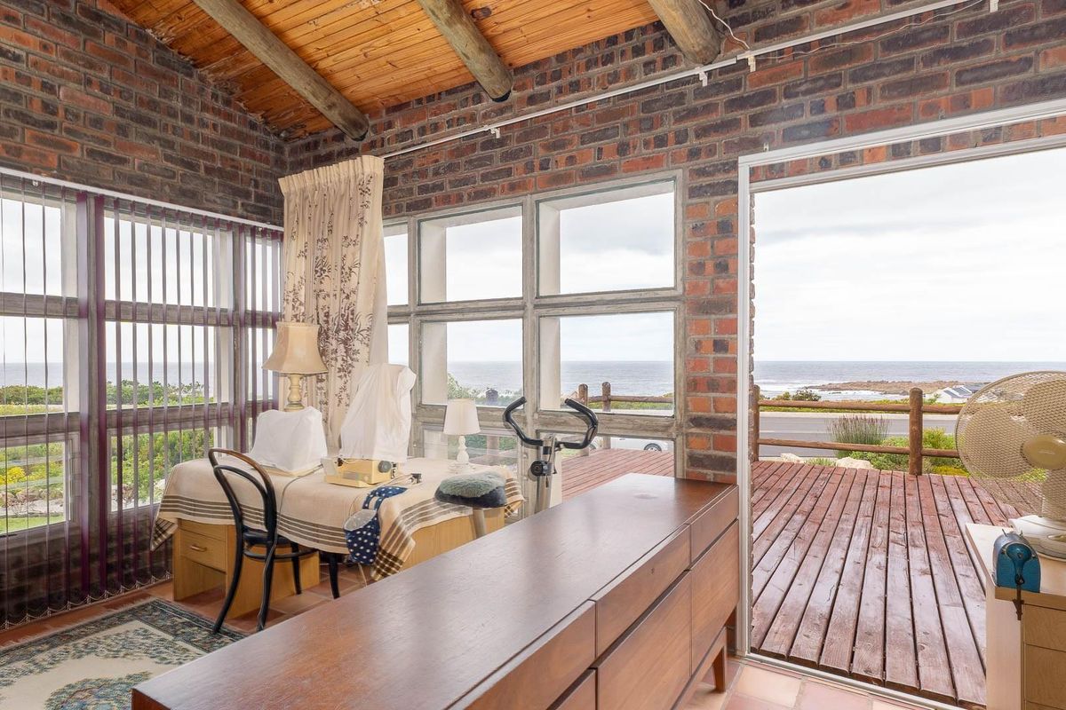 A living area featuring exposed brick walls, timber beamed ceiling, and large windows with direct access to an outdoor wooden deck overlooking the ocean.
