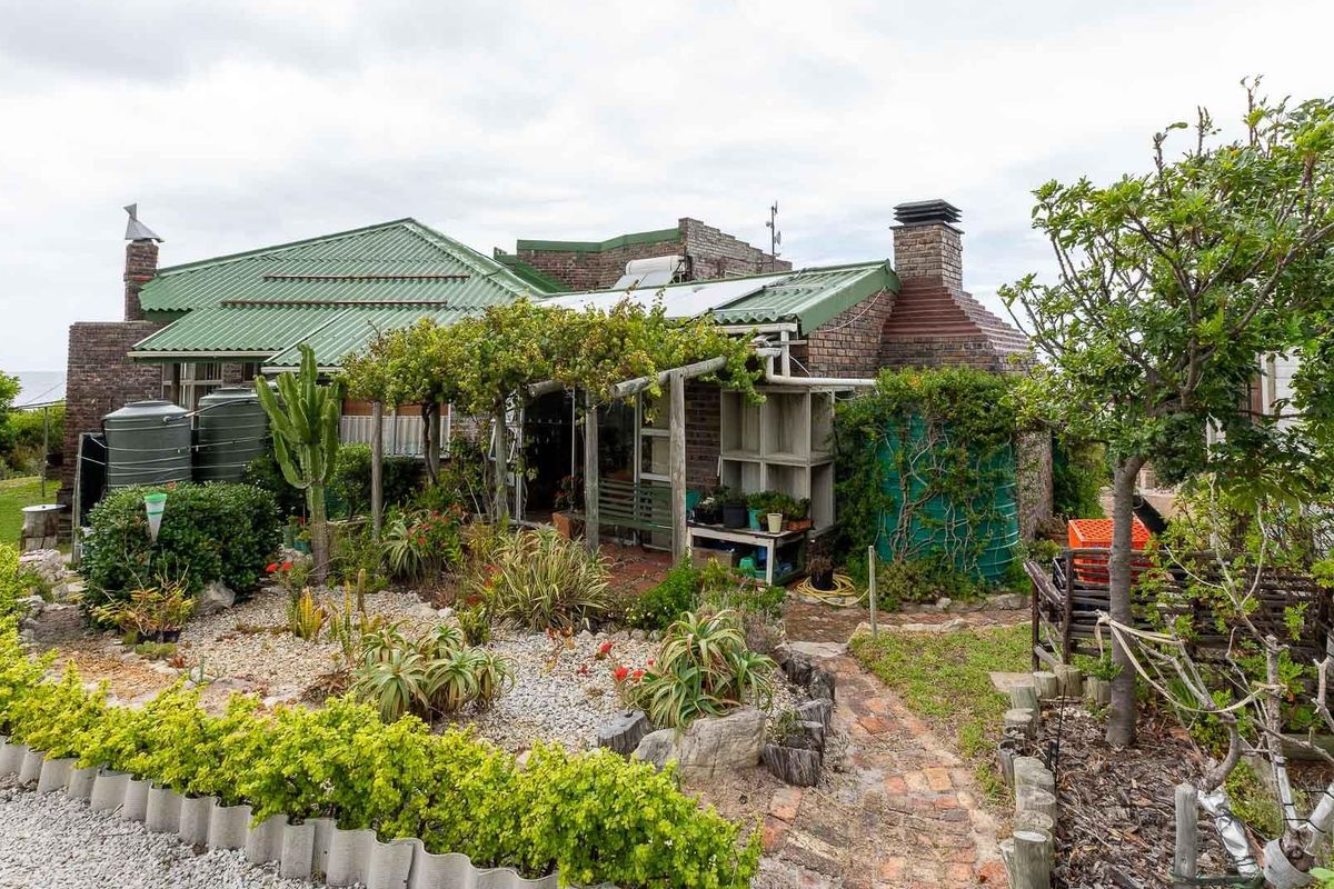 A rear view of a brick house featuring a green metal roof, a timber pergola, and a landscaped garden with water storage tanks.