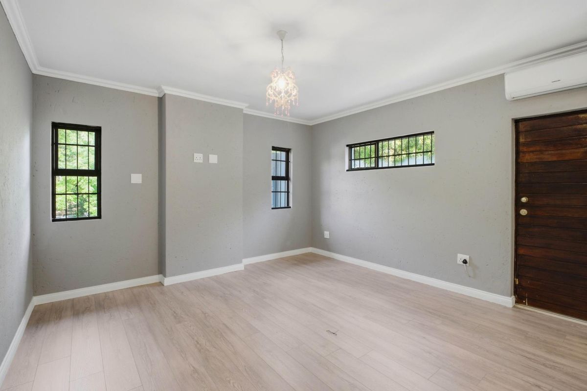 A bright, empty living room featuring light wood-look flooring, grey textured walls, black-framed windows, and a wall-mounted air conditioning unit.
