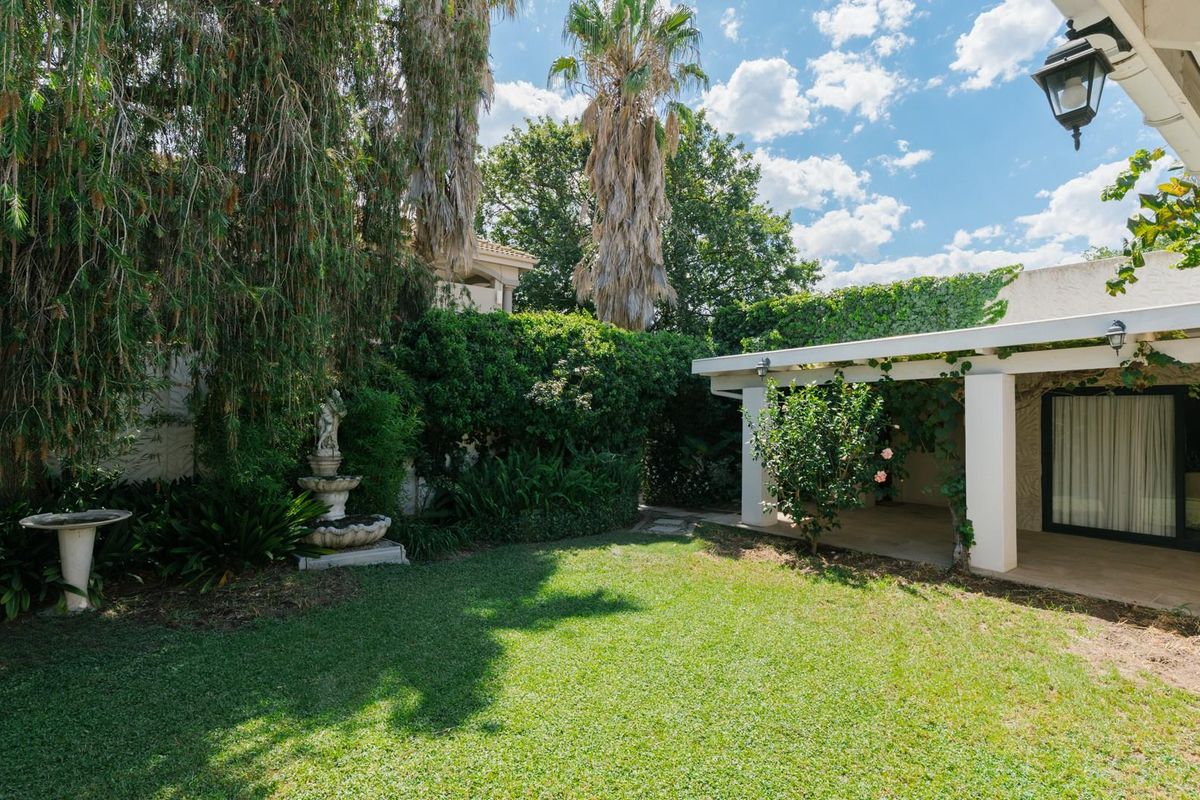 A well-maintained rear garden featuring a covered patio area, lush landscaping, mature trees, and a decorative stone fountain.
