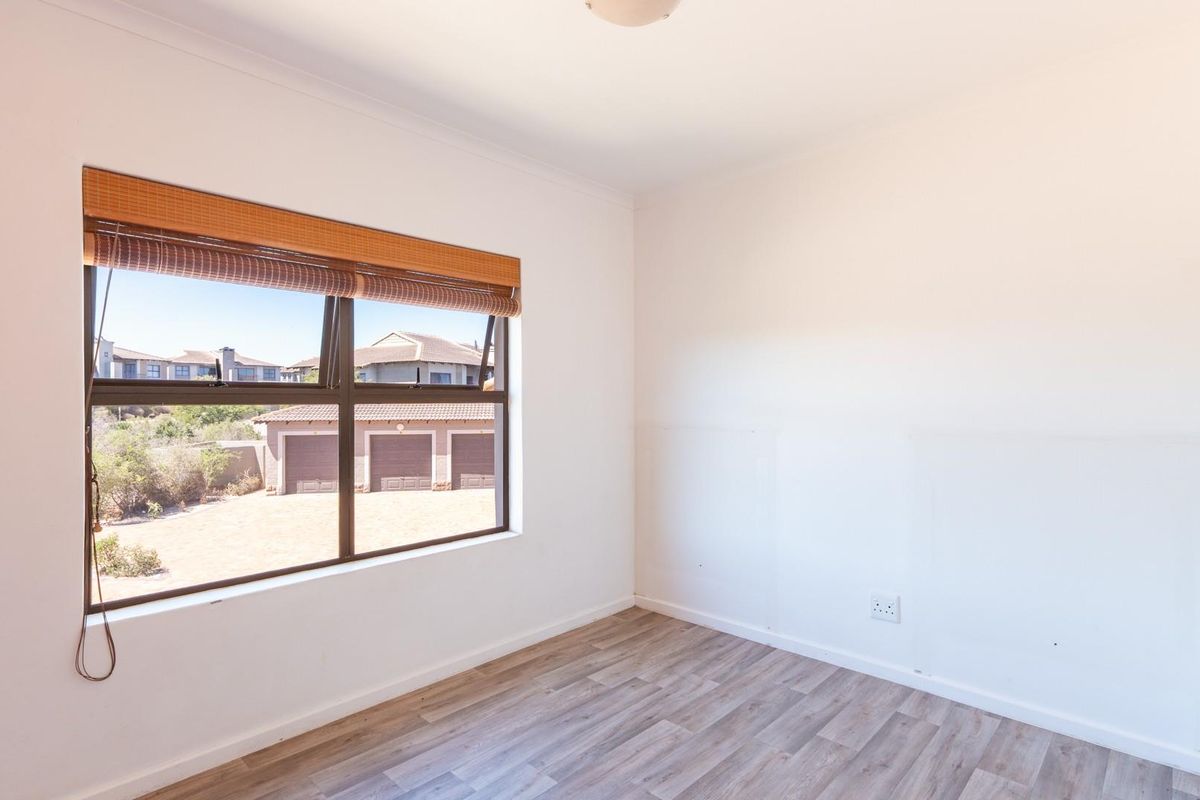 A bright, empty bedroom featuring modern wood-look laminate flooring, a large window with a roller blind, and neutral white walls.