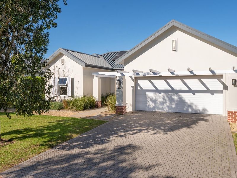 A well-maintained single-story house featuring a paved driveway, double garage, and a decorative pergola, set within a neat landscaped garden.