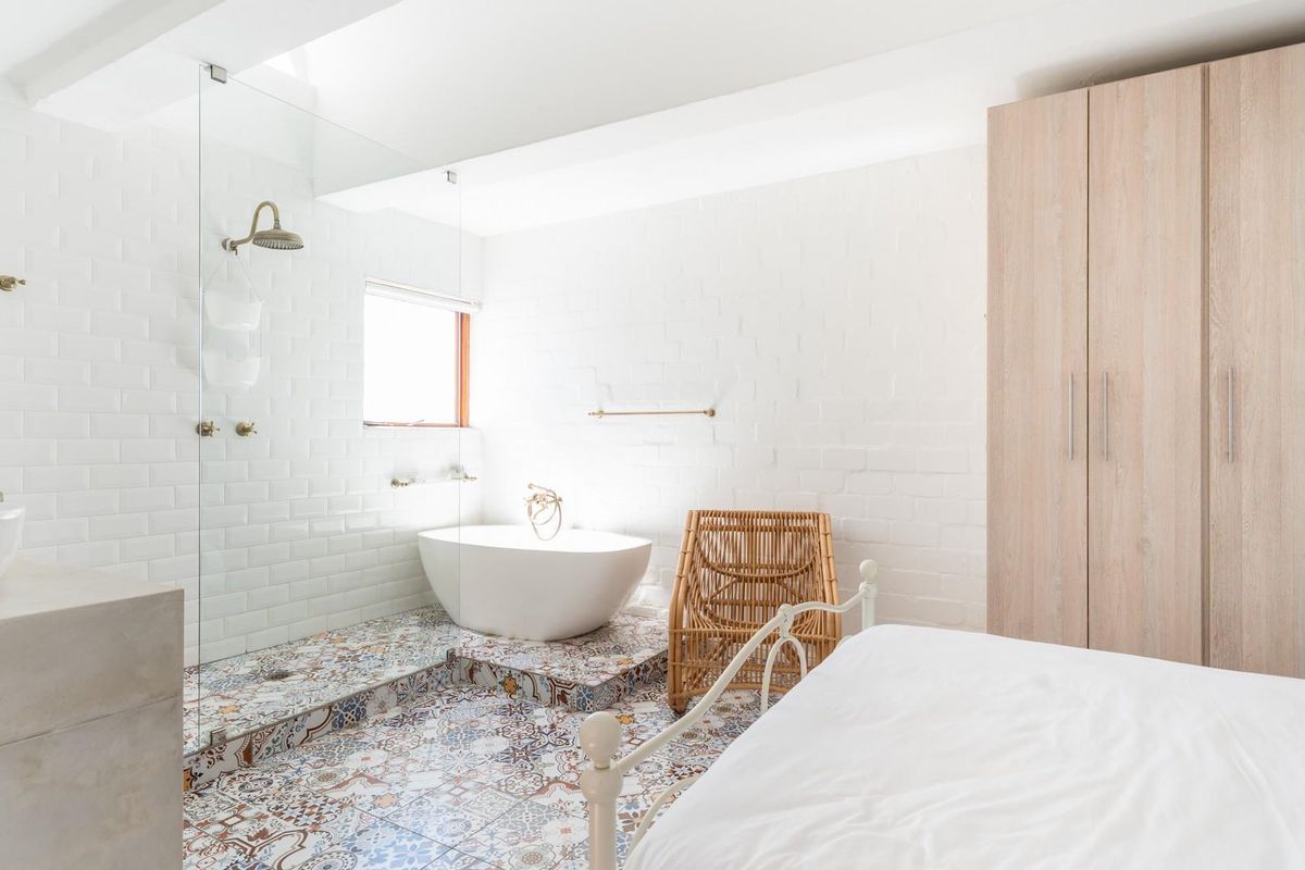 A bright, modern bedroom featuring an integrated open-plan bathroom with a freestanding bathtub, glass-enclosed shower, patterned tile flooring, and built-in wooden cabinetry.