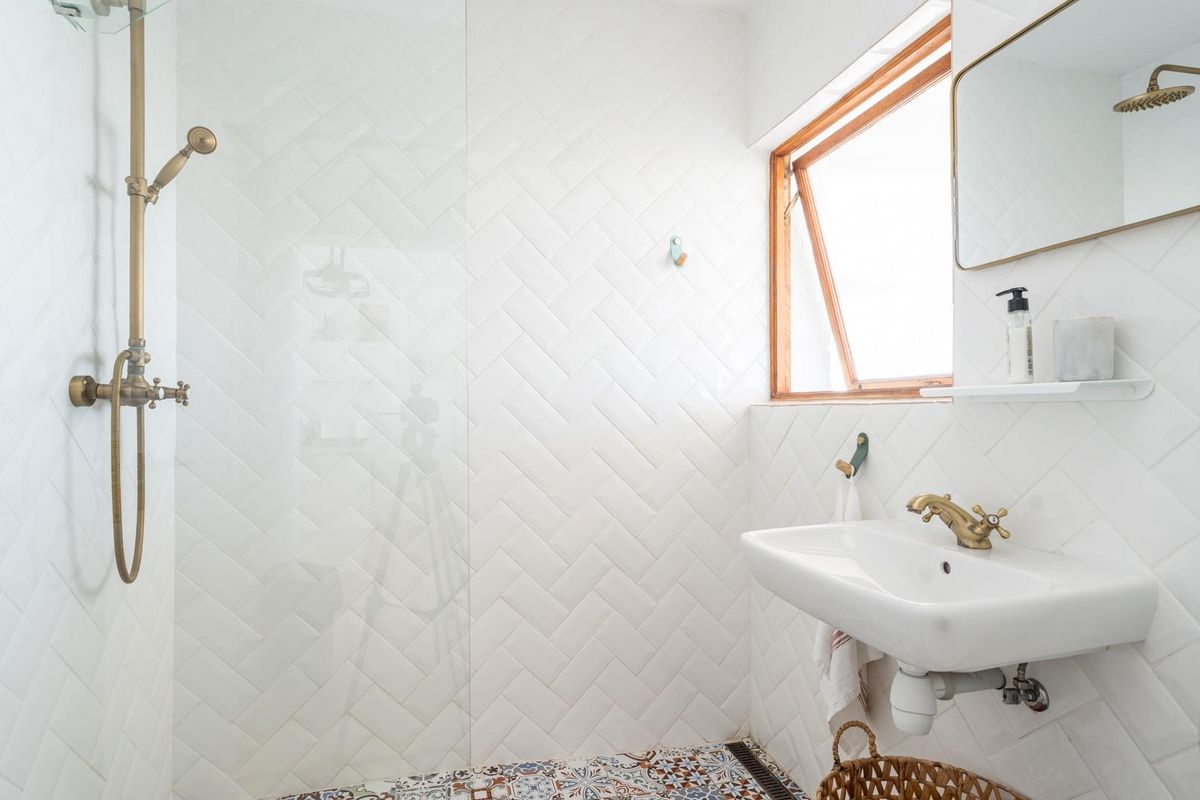 Modern bathroom featuring a walk-in shower with brass fixtures, white herringbone wall tiles, patterned floor tiles, and a wall-mounted basin.