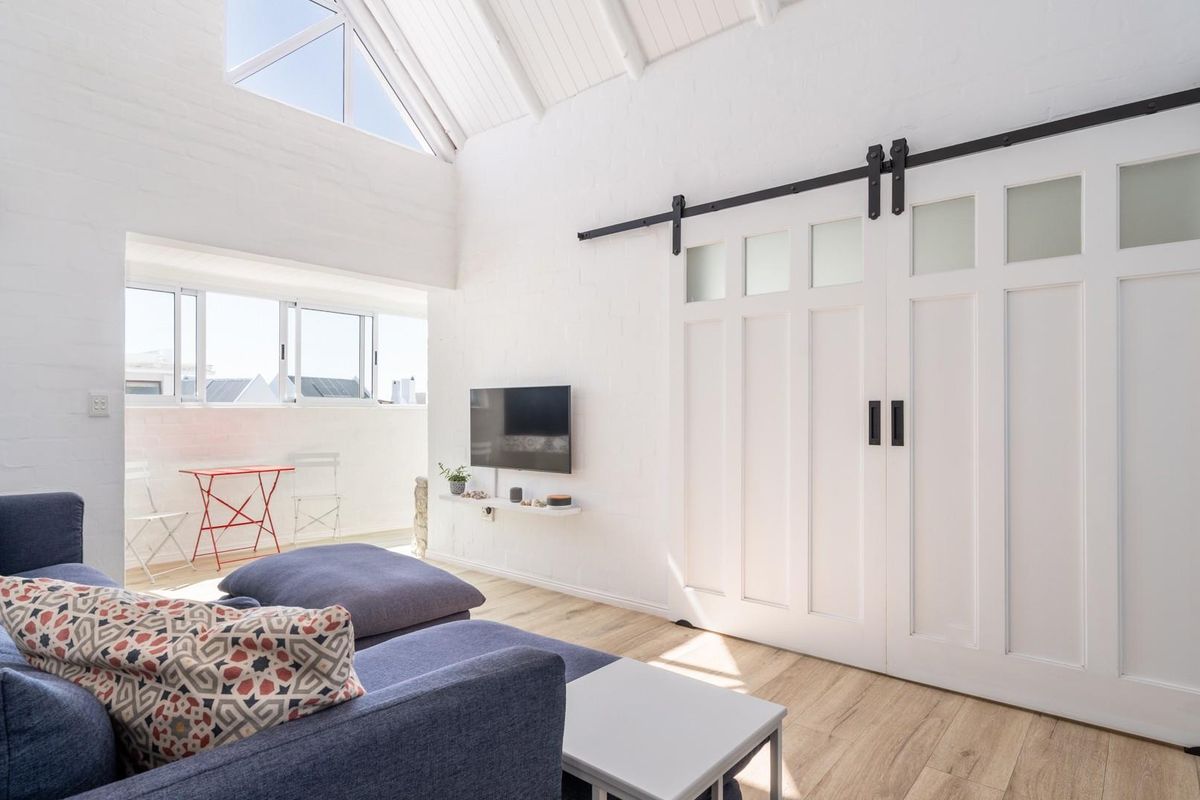 Bright living area featuring high vaulted ceilings, exposed white brick walls, wood-look flooring, and large sliding barn doors leading to an adjoining space.