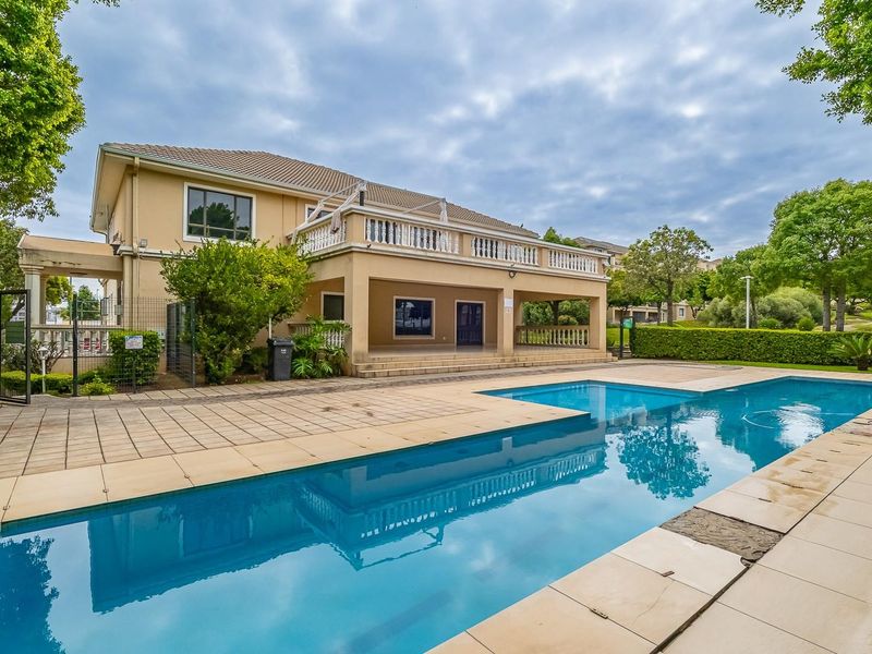 A well-maintained communal swimming pool area featuring a paved deck, landscaped garden surrounds, and a covered patio area attached to the apartment building.