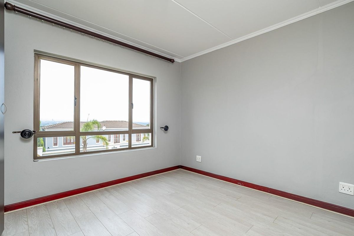 A bright, empty bedroom featuring light-toned laminate flooring, neutral grey walls, and a large window with a curtain rod.