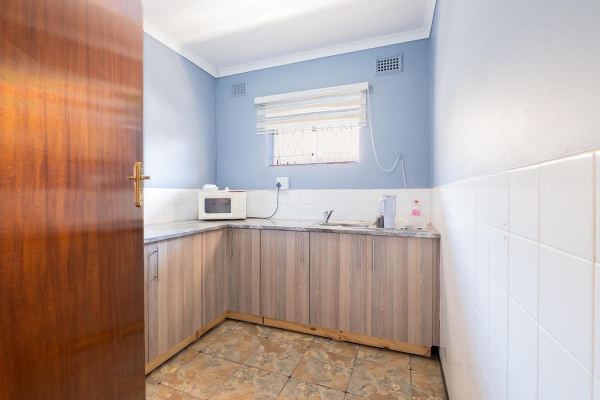A compact kitchenette featuring wood-look cabinetry, laminate countertops, a stainless steel sink, and tiled splashbacks in a functional office space.