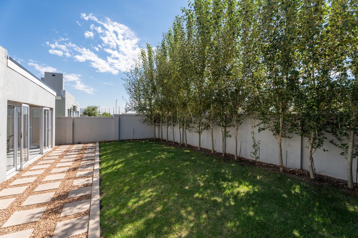 A well-maintained rear garden featuring a paved walkway, manicured lawn, and a perimeter wall lined with mature trees.