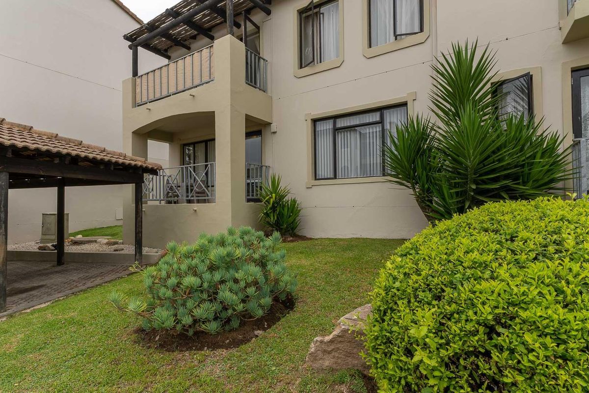 Exterior view of the apartment building featuring a covered patio, balcony, and a well-maintained garden area with a paved carport structure.