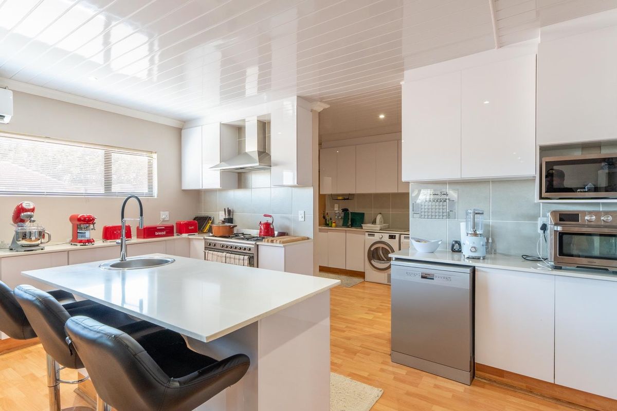 Modern kitchen featuring white cabinetry, stone countertops, a central island with sink, integrated oven, and a separate scullery area with laundry facilities.