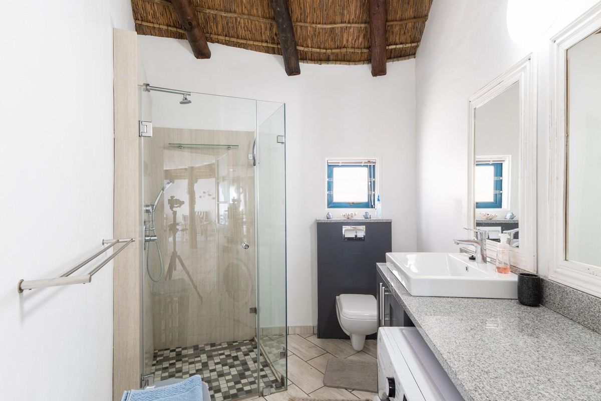 A modern bathroom featuring a glass-enclosed shower, wall-mounted toilet, granite vanity countertop with a basin, and a unique exposed thatch roof ceiling.