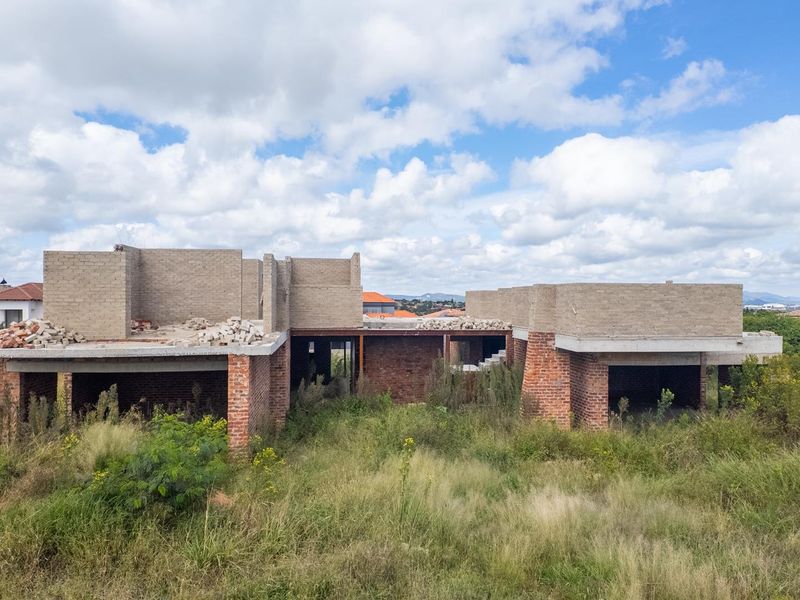 An aerial view of a residential property under construction, featuring exposed brick walls and concrete structural elements on a large, overgrown plot.