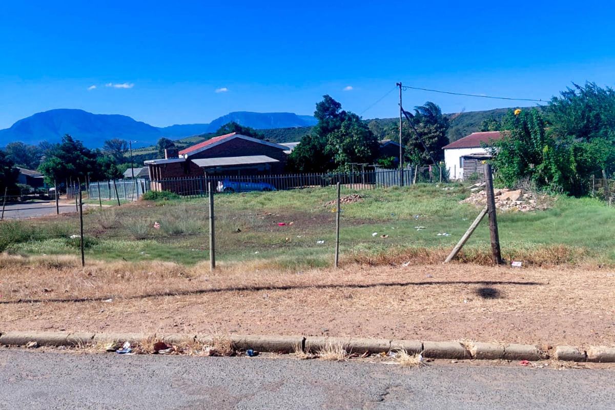 A vacant residential plot featuring a wire fence, utility pole, and street-side kerb, offering a clear view of the surrounding neighbourhood and distant mountains.