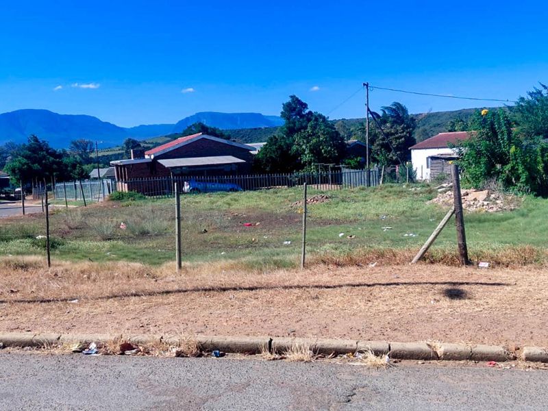 A vacant residential plot featuring a wire fence, utility pole, and street-side kerb, offering a clear view of the surrounding neighbourhood and distant mountains.