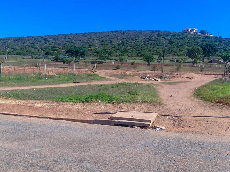 A vacant residential plot featuring road frontage, wire fencing, and a concrete stormwater drain cover, set against a hilly landscape.