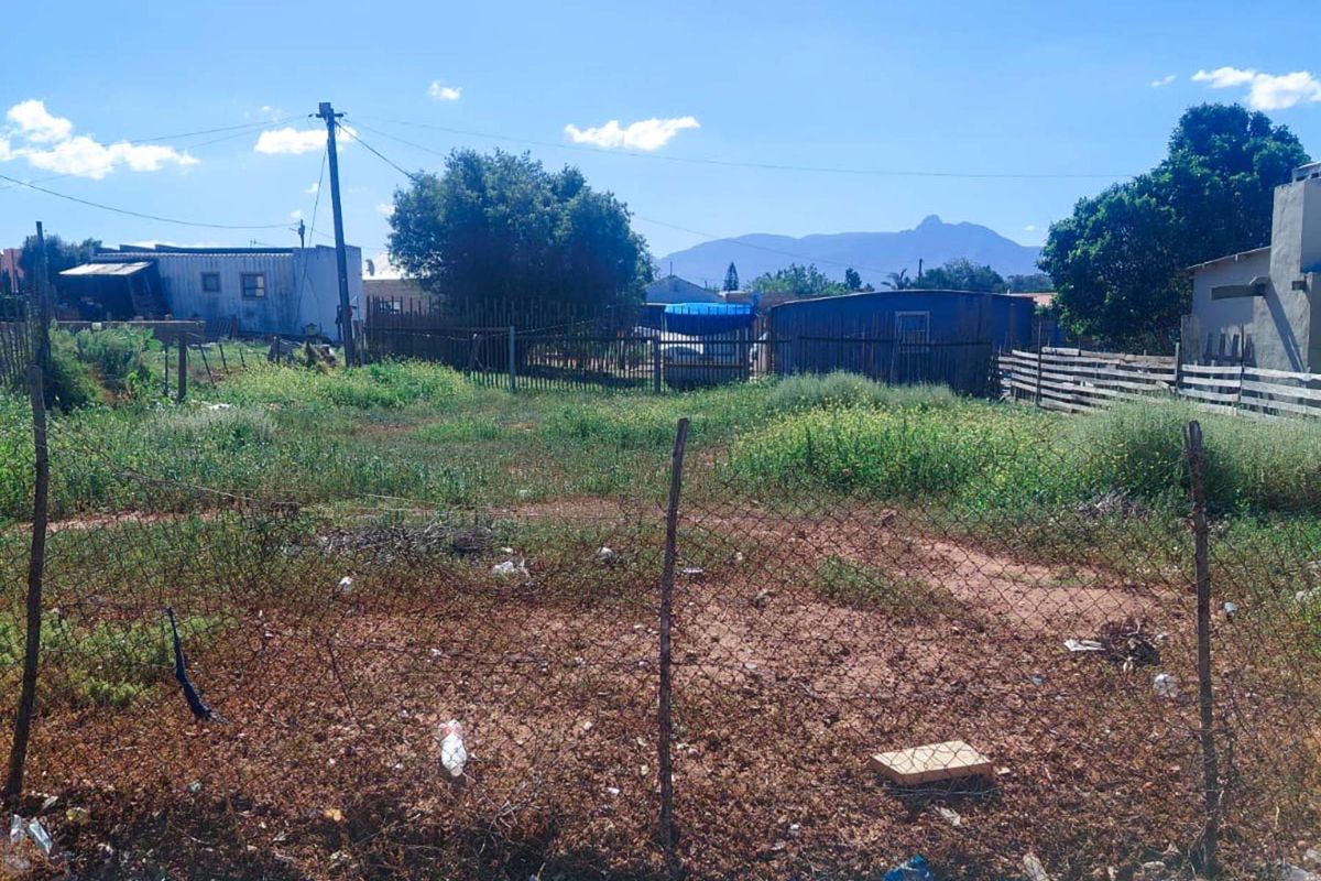 A vacant residential plot featuring wire mesh fencing and a utility pole, with surrounding neighborhood structures and mountain views in the distance.